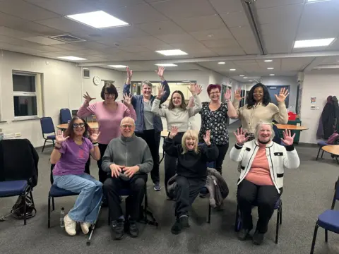 A group of nine people, four seated in front of nine standing, all waving their hands in the air and smiling.
