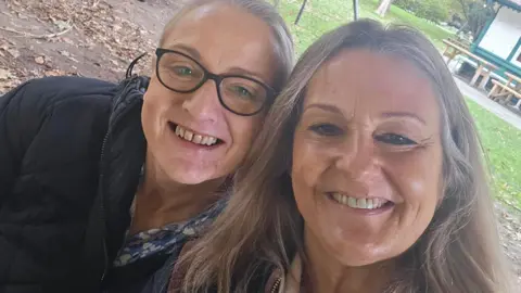 Submitted A selfie of Nicola Kilcoyne and Vanessa Davidson standing in a park. They both have long light brown hair which is slightly greying, and Ms Kilcoyne is wearing dark framed glasses. There are benches in the background.