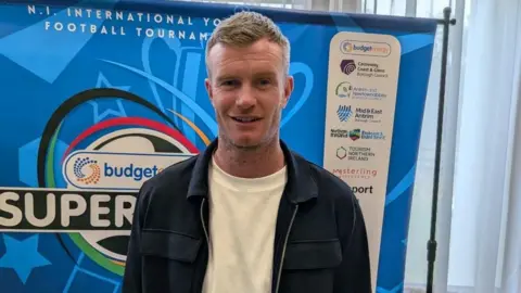 Chris Brunt has short brown hair and is wearing a navy jacket and white shirt. He is standing in front of a board with a number of sponsors.