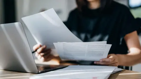 Cropped shot of an anonymous woman holding banking and finance papers with laptop on the table in front of her. 