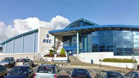 The exterior of the National Sports Centre, a large blue building with arching rooves. 