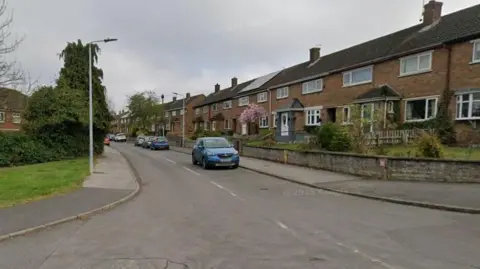 Google Road with houses on both sides. There are a few cars.