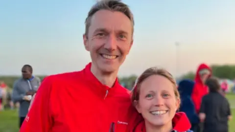 Mike Green stands with his arms around his wife Collette. They are both smiling at the camera. They are standing outside in a sports pitch area with people behind them. Mike has short light hair and wears a red running jumper. Collette has light hair and wears a red hoodie with a coat over the top.