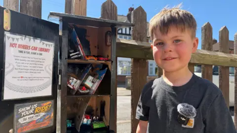 A young boy smiles to camera wearing a grey t-shirt. To his left is a birdbox style toy swap box full of toy cars.
