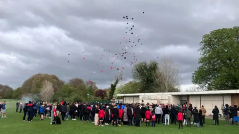 Sam Read/BBC Hundreds of people stand in a football field and watch as tens of colourful balloons float above them into the sky on a grey day.