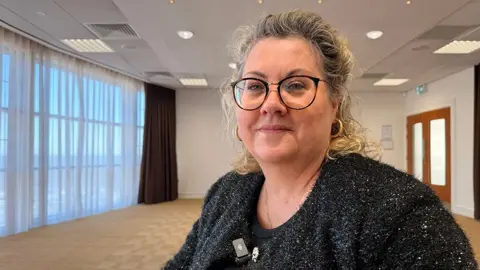 A picture of Lisa Squire. She has short blond hair and wears glasses and a black top. She is standing in an empty room and looking into the camera.