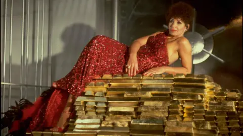 Getty Images The singer Dame Shirley Bassey, pictured in the 1980s. She is wearing a sparkly red dress and lying on a large pile made up of bars of gold.