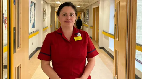 Louise Proudfoot, a woman with brown hair, wearing in a red matron uniform in a hospital corridor.