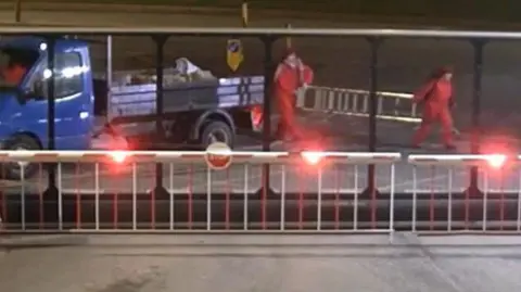 Still from CCTV shows two women in red overalls carrying a silver ladder behind a large security fence. Their associate is sitting in the cab of a blue flatbed truck next to them with a pile of rubble on its back.