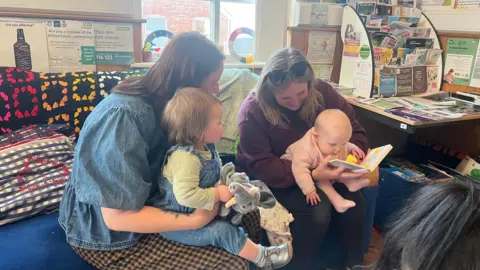 Two mums in a community centre, sitting on a sofa. Both are holding a baby, one of whom is looking closely at a book. The other child is holding a stuffed elephant toy.