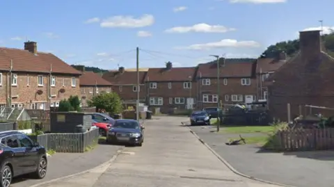 Google Brown brick houses in an Esh Winning street. There is a bike lying on its side and a number of parked cars. 