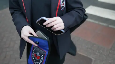 A student in a dark school uniform with red piping is placing a smartphone into a school branded pouch