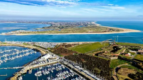 kevdrone An aerial photo shows the shoreline at Eastney with yachts moored in the harbour. In the distance you can see the Hayling Island peninsular with golden sands. The sea is as deep blue. 