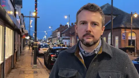 Pallion ward councillor Martin Haswell standing on Saint Luke's Terrace in Pallion with street lights and shops in the background. He has a grey coat and a beard and moustache.