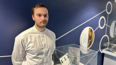 A man with brown hair and stubble standing beside a glass display case inside. He is wearing a white shirt.