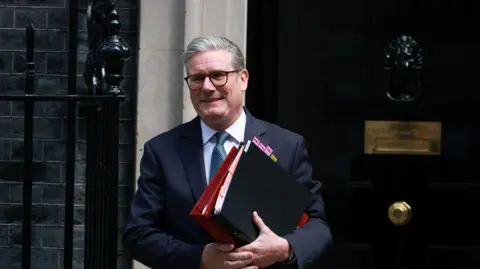 PM Keir Starmer walking out of 10 Downing st. He has two large ring binder folders in his hands and is wearing a dark blue suit with light blue tie
