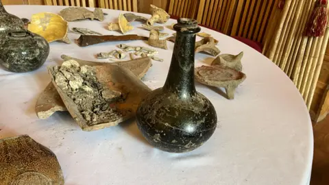 Henry Godfrey-Evans/BBC A mix of artefacts, broken pottery and bones laid out on a table with a white sheet on it.