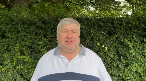 Councillor John Hubbard stands in front of a hedge while wearing a light grey shirt. He has short grey hair and grey stubble.