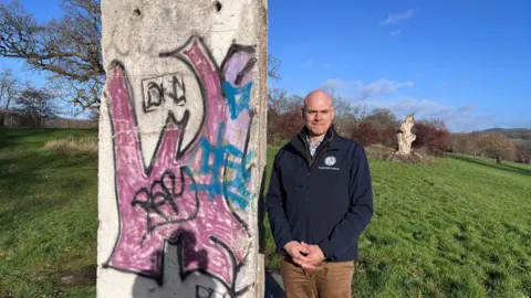 A bald man in blue jacket and brown trousers next to a piece of the Berlin Wall