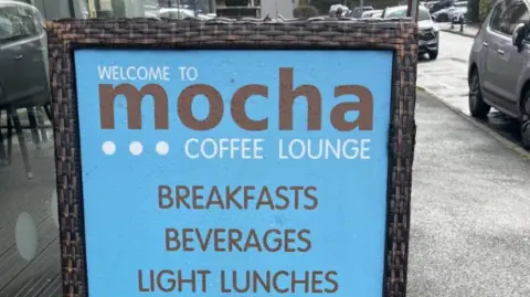 Blue sign post from outside a cafe. It says Welcome to Mocha Coffe Lounge Breakfasts Beverages Light Lunches.