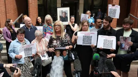 PA A group of people stand outside a redbrick public building, surrounded by microphones, cameras and mobile phones. Several participants hold printed signs reading: “REGULATE OUR FUNERAL INDUSTRY” with large QR codes, while others hold framed and printed photographs of older adults. In the centre, a person speaks while holding a framed photograph, with supporters gathered closely around. The building entrance and steps form the backdrop.