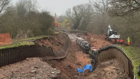 Two narrowboats sit in a water-less canal surrounded by mud and collapse banking. People in fluorescent jackets stand on the embankment by machinery with further machinery visible in the distance. 