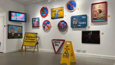 A wide picture of a white art gallery wall covered with roadworks signage and paintings.
