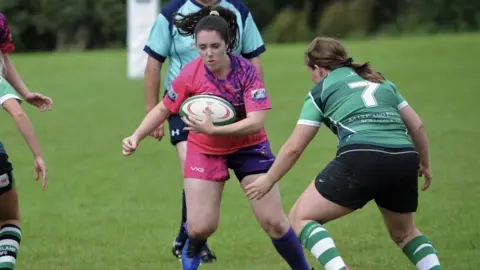 Ken Langley A young brunette girl dressed in a pink kit, holding a rugby ball whilst trying to dodge an opponent. 
