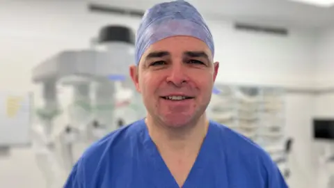 A head and shoulders photo of Dr Burton looking directly at the camera. He is wearing a blue, v-necked surgical gown and a surgical cap on his head.  
