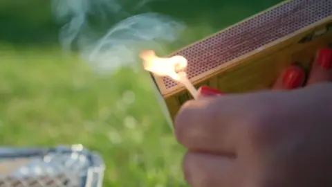 BBC Close up of a hand striking a match against a matchbox. In the background the corner of a disposable barbecue is positioned on some grass.