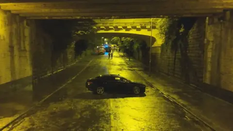 Merseyside Fire and Rescue Service A black car on a stretch of road underneath a railway bridge, with a fire engine parked in the background.