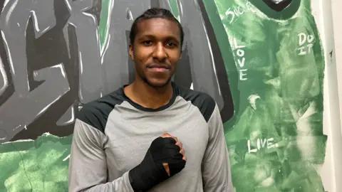 Philip stands in front of a wall with green and black graffiti on it. He is wearing a grey and black long-sleeved T-shirt and his right hand is clenched across his chest, it has black strapping on it. He has black cornrows. 