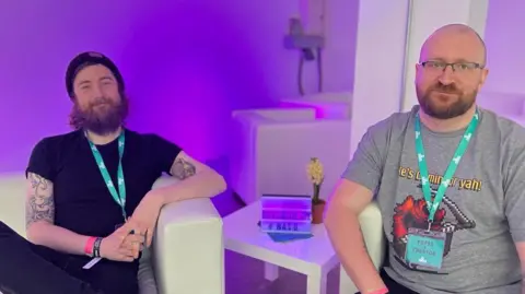 ANDY COX Two men - both bearded and wearing turquoise lanyards are sat in white armchairs in front of a purple-lit background. 