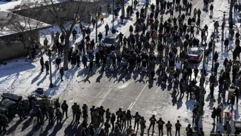 An aerial image that shows a line of about 20 ICE agents on one side of the road facing about 50 protesters on the snowy street.