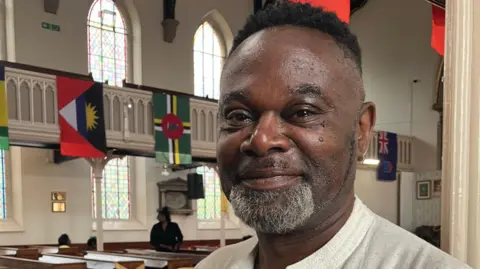BBC/Phil Bodmer A man stands inside a church. Flags from around the country can be seen hanging in the background as well as rows of wooden pews.