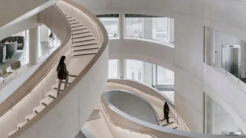 Royal Institute of British Architects Winding, white concrete staircase. One person is walking up one and another is in the distance walking up another staircase. The building's interior also has a concrete-look and is white. 