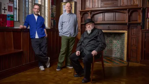 Royal Shakespeare Company George R.R. Martin is seated and wearing a black shirt over black trousers. He is also wearing a black bowler hat. On the left of the image is the play's write Duncan Macmillan in a bright blue shirt over a white t-shirt, dark trousers and white trainers. Between them is the director Dominic Cooke, who is wearing a light blue-grey shirt over olive drab trousers and dark trainers. they are in a dark wood panelled room.