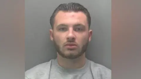 Cambridgeshire Police A police custody photo of Chas Corrigan. He has short brow hair and beard and is wearing a grey jumper. He is standing in front of a grey wall.
