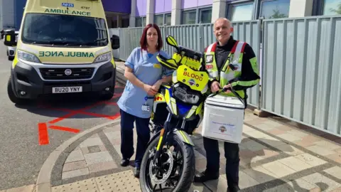 DERBYSHIRE BLOOD BIKES Adam, a volunteer blood biker, stands next to his motorcycle with a nurse outside Royal Derby hospital. He is dressed in neon motorcycle kit and holding a medical courier bag of breast milk. Behind them, there is an ambulance parked.