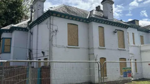 A white-walled building with boarded up windows and a dark grey roof with metal fencing surrounding it