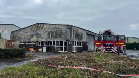 Image shows partially collapsed warehouse at a Plympton business park. The building is partially collapsed. A fire engine is parked next to the building. 