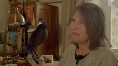 Sara Cox sits at her desk. She has shoulder length dark hair with a pair of glasses on top of her head. Behind her sits a large black raven made from wire