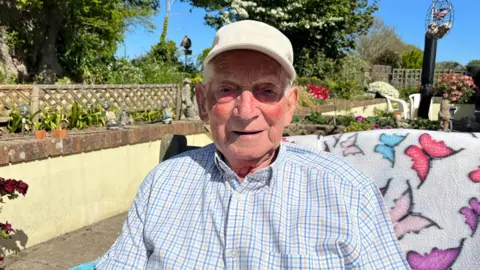 A man with white hair and a beige cap is looking into the camera. He has white, blue and yellow checked shirt on. He's sitting on a patio on a wooden bench and there are raised flower bed behind him , as well as a bird feeder. It's sunny and his face is shaded slightly by his hat.