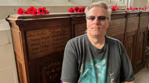BBC/Lucy Ashton A man with short grey hair and glasses is wearing a t-shirt. He is sitting in a church foyer in front of a wooden shelf with poppy wreaths