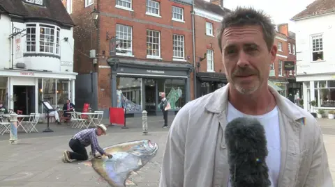 Jim Murray, look at the camera, with a large chalk salmon being drawn on the pavement behind him by the artist Julian Beever, who can be seen on his knees.