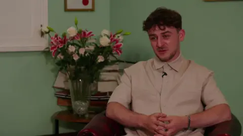 Gersi sits in a chair and looks off camera towards a reporter during a TV interview. He is wearing a beige shirt and has brown hair