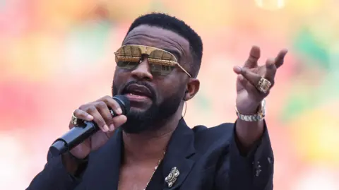 Papa Wemba sings on stage wearing a black suit and large encrusted jewellery. In one hand he holds a microphone and with the other he reaches out.