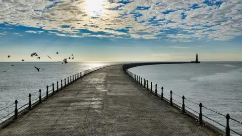 BBC Weather Watchers/Richard R Clark Roker Pier which is curved with railings on either side. In the distance is the lighthouse which is just a silhouette. A flock of birds are flying close to the pier on the left. The sky is blue and white with small, wispy clouds in front of the sun.