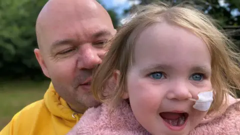 Handout A family photo showing Terry, wearing a yellow hoody and holding up his daughter Beatrix, who has blonde hair, blue eyes and is wearing a fluffy pink jacket. They are both in a park with trees in the background.