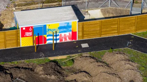 Marcin Jedrysiak A wooden fence with a coloured block with the words 'Female showers and toilet' with old pictures and colourful designs